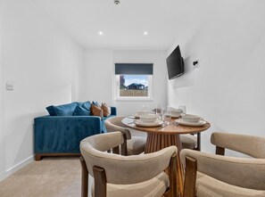 Ein modernes Wohnzimmer mit Esstisch für vier Personen, einem blauen Sofa und einem an der Wand montierten Fernseher.