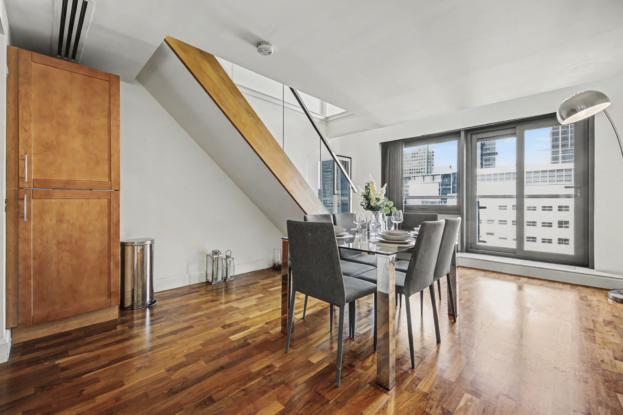 Un comedor moderno con una escalera de madera, una mesa para cuatro personas y grandes ventanales con vista a la ciudad.