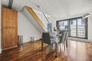 Uma área de refeições moderna com escada de madeira, mesa posta para quatro pessoas e grandes janelas com vista para a cidade.