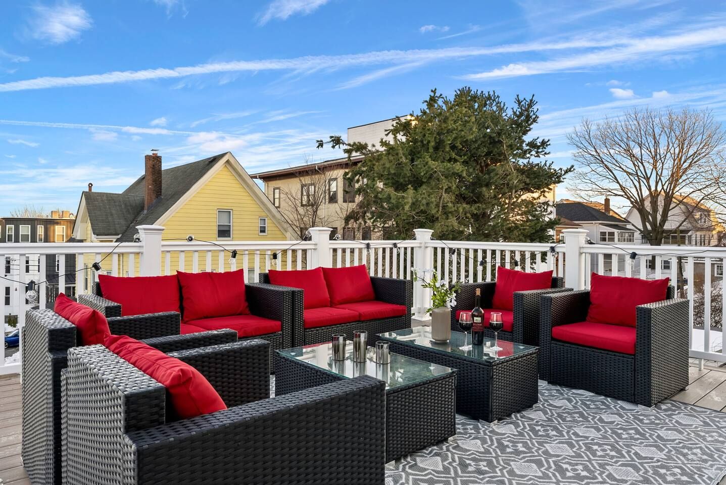 Une terrasse dotée de meubles en osier et de coussins rouges, d’une table en verre et offrant une vue sur les maisons au loin.