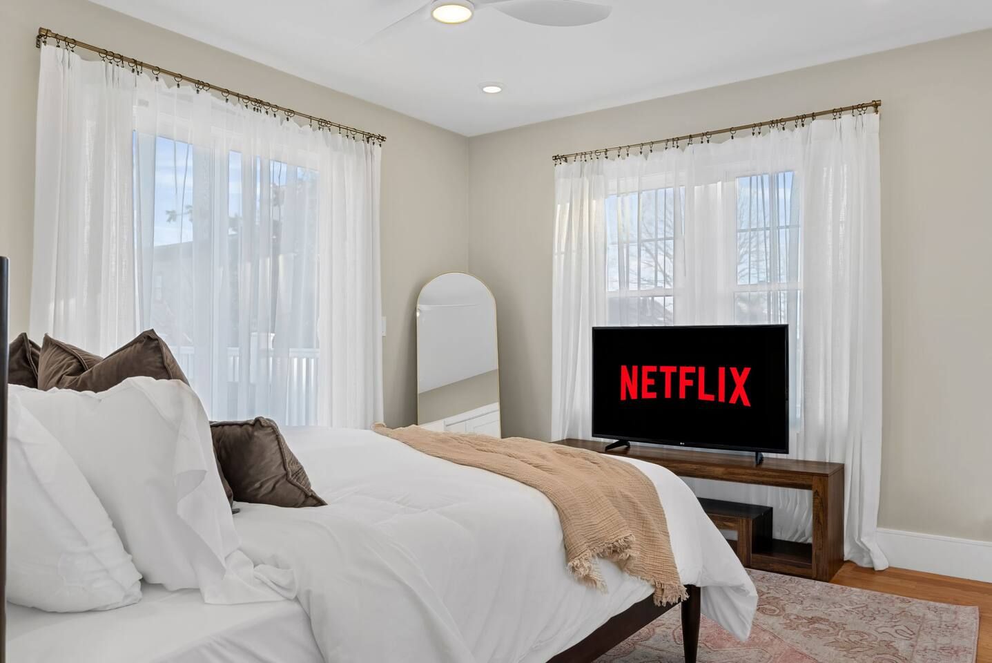 Une chambre à coucher avec un lit, un téléviseur affichant le logo « NETFLIX », et une fenêtre ornée de rideaux légers.