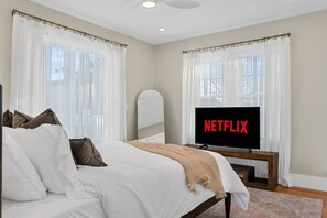 Une chambre à coucher avec un lit, un téléviseur affichant le logo « NETFLIX », et une fenêtre ornée de rideaux légers.