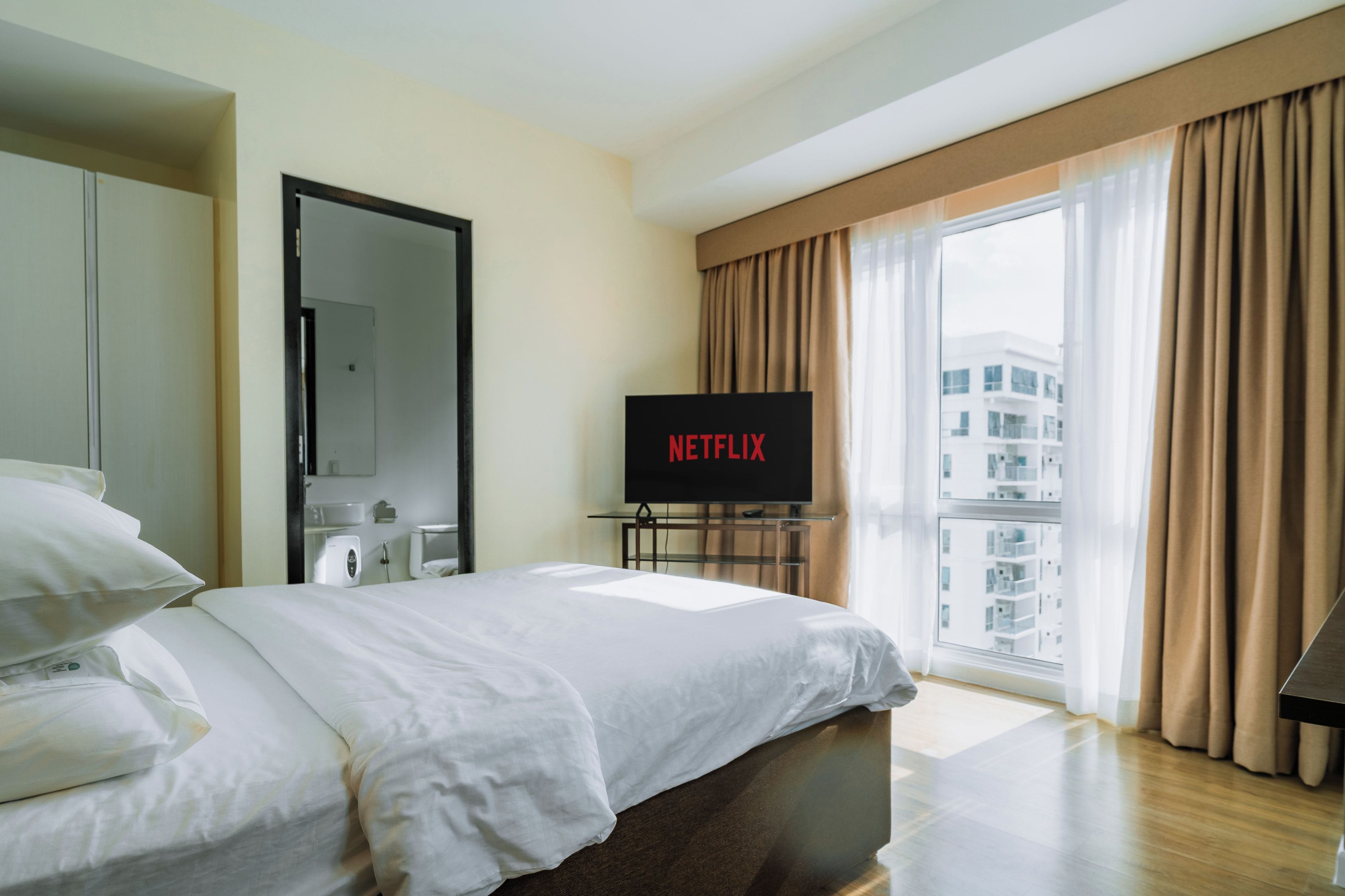 Habitació d'hotel amb un llit, una televisió que mostra 'NETFLIX', un bany amb mirall i una finestra amb cortines.