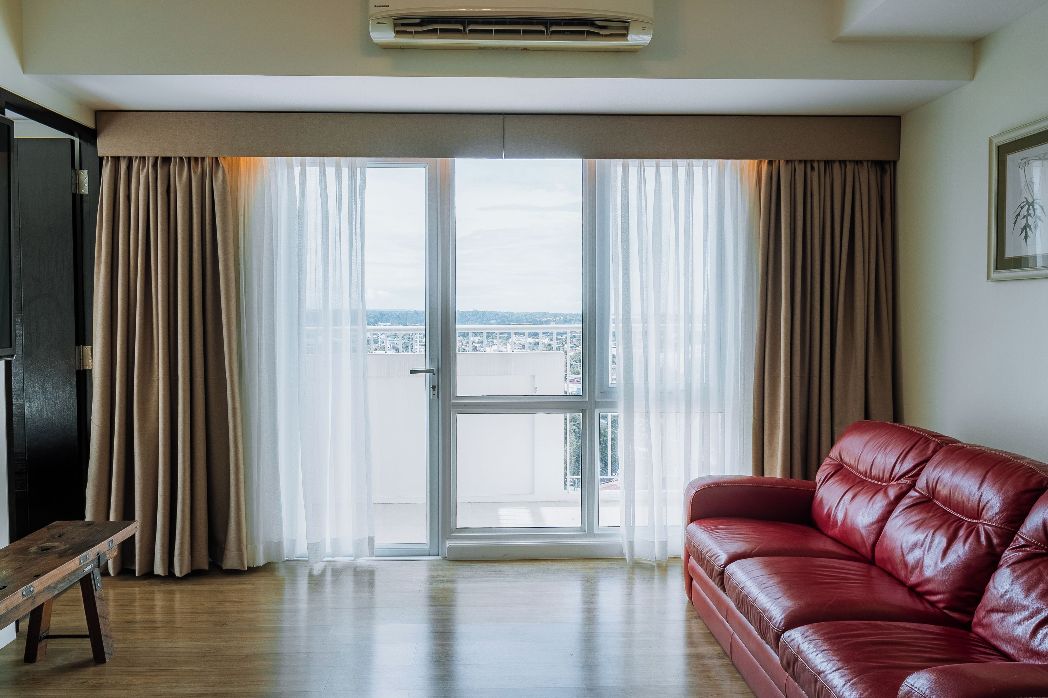 A room with a red leather sofa, a wooden bench, and a large sliding glass door leading to a balcony with a view of a bridge and buildings.