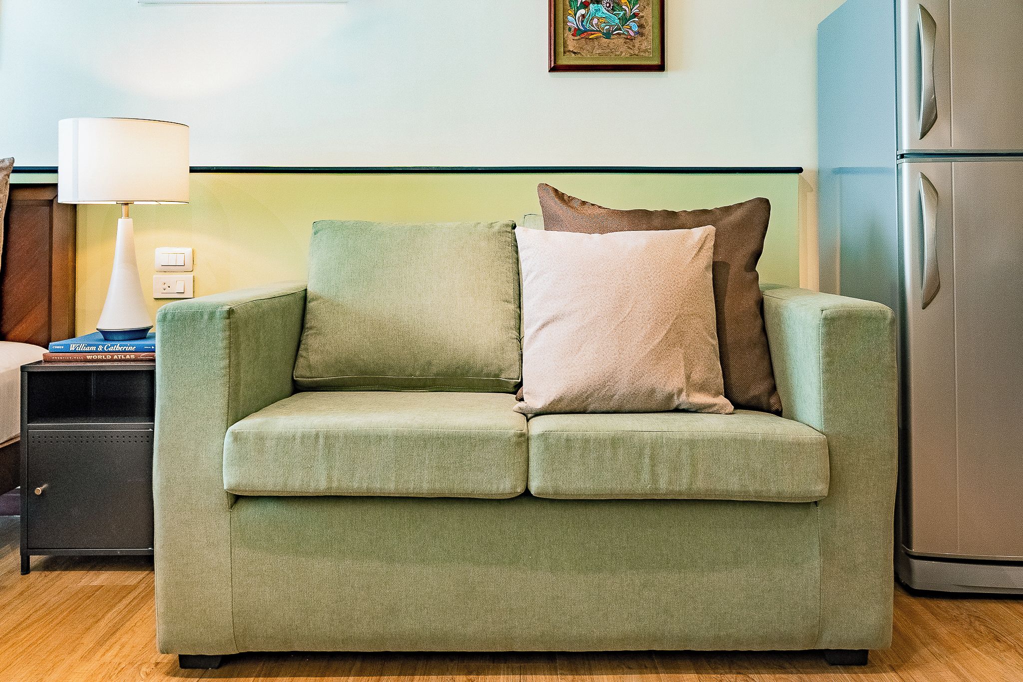 A green sofa with two pillows, a small table with a lamp, and a refrigerator in a room with a light green wall and a framed picture.