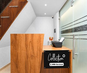 A reception desk with a wooden finish, a black sign reading 'Collection' and 'YOO-Serviced', and a glass partition to the right. - Classic Hotel Shreeda (Greater Noida)
