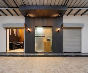 A modern building entrance with a glass door, a wooden interior visible through the door, and a closed roller shutter on the right. - Classic Hotel Shreeda (Greater Noida)