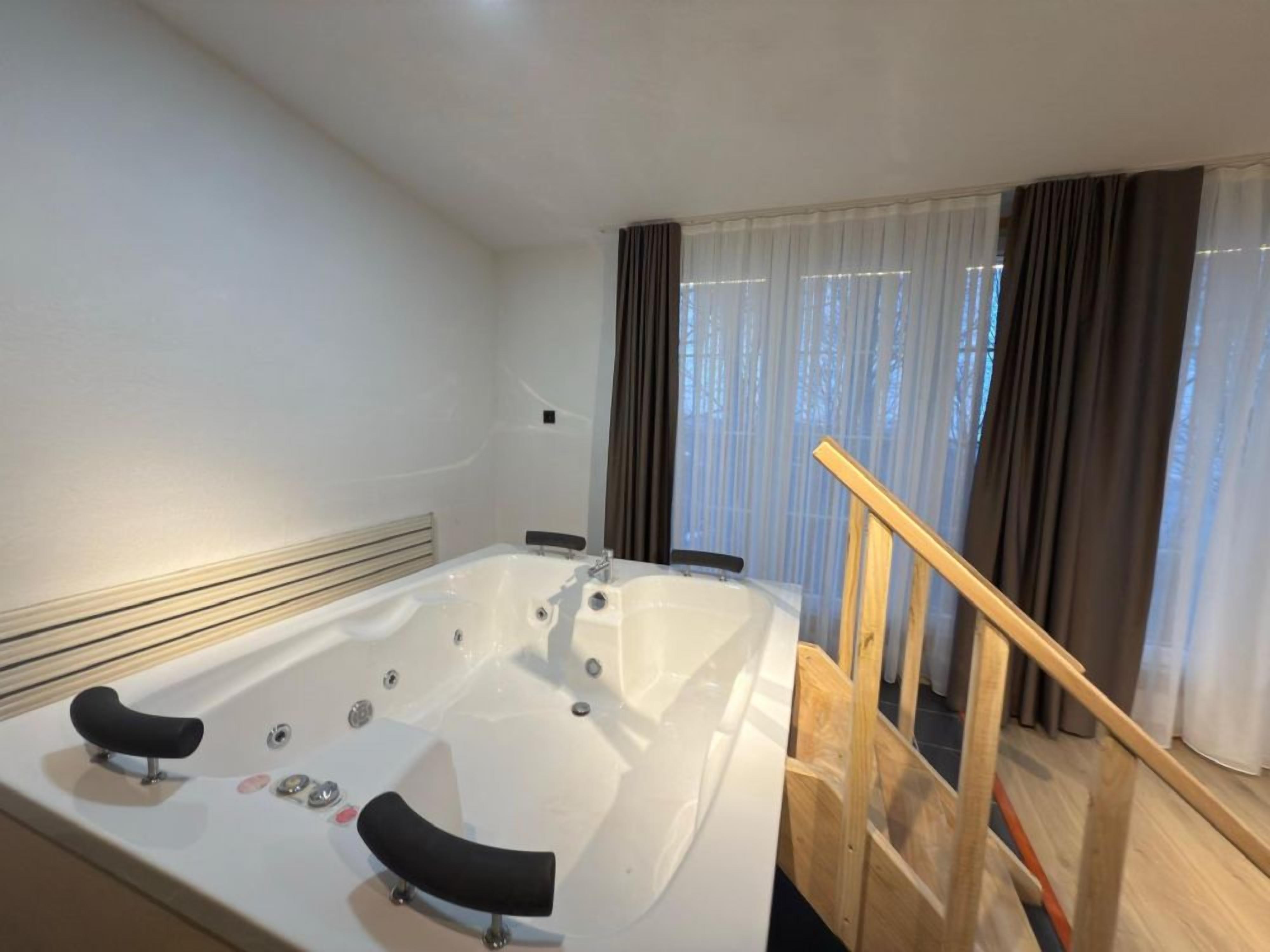 A modern bathroom with a large bathtub, wooden staircase, and a view of the outdoors through sheer curtains.
