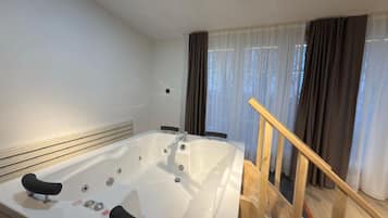 A modern bathroom with a large bathtub, wooden staircase, and a view of the outdoors through sheer curtains.