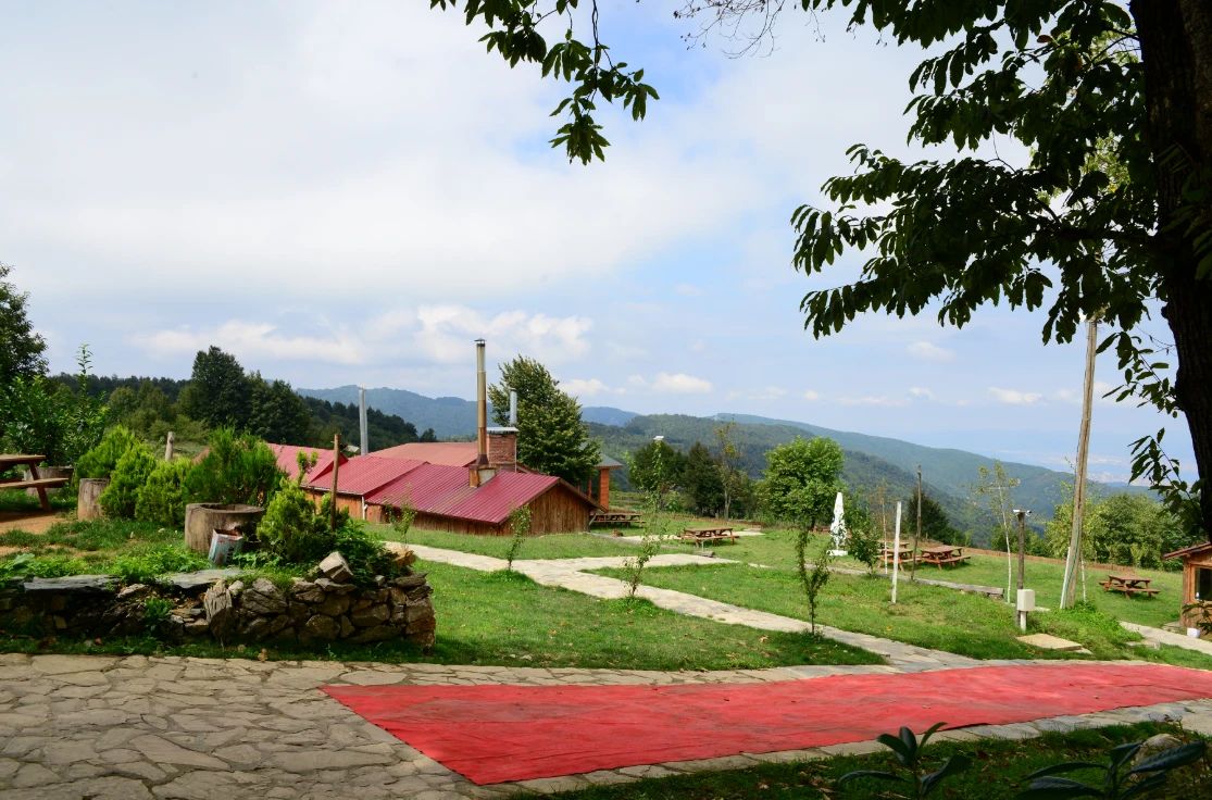 Un edificio rústico con techo rojo, rodeado por un muro de piedra, una alfombra roja y mesas de picnic en un área cubierta de césped, con montañas al fondo.