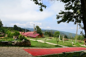 Un edificio rústico con techo rojo, rodeado por un muro de piedra, una alfombra roja y mesas de picnic en un área cubierta de césped, con montañas al fondo.