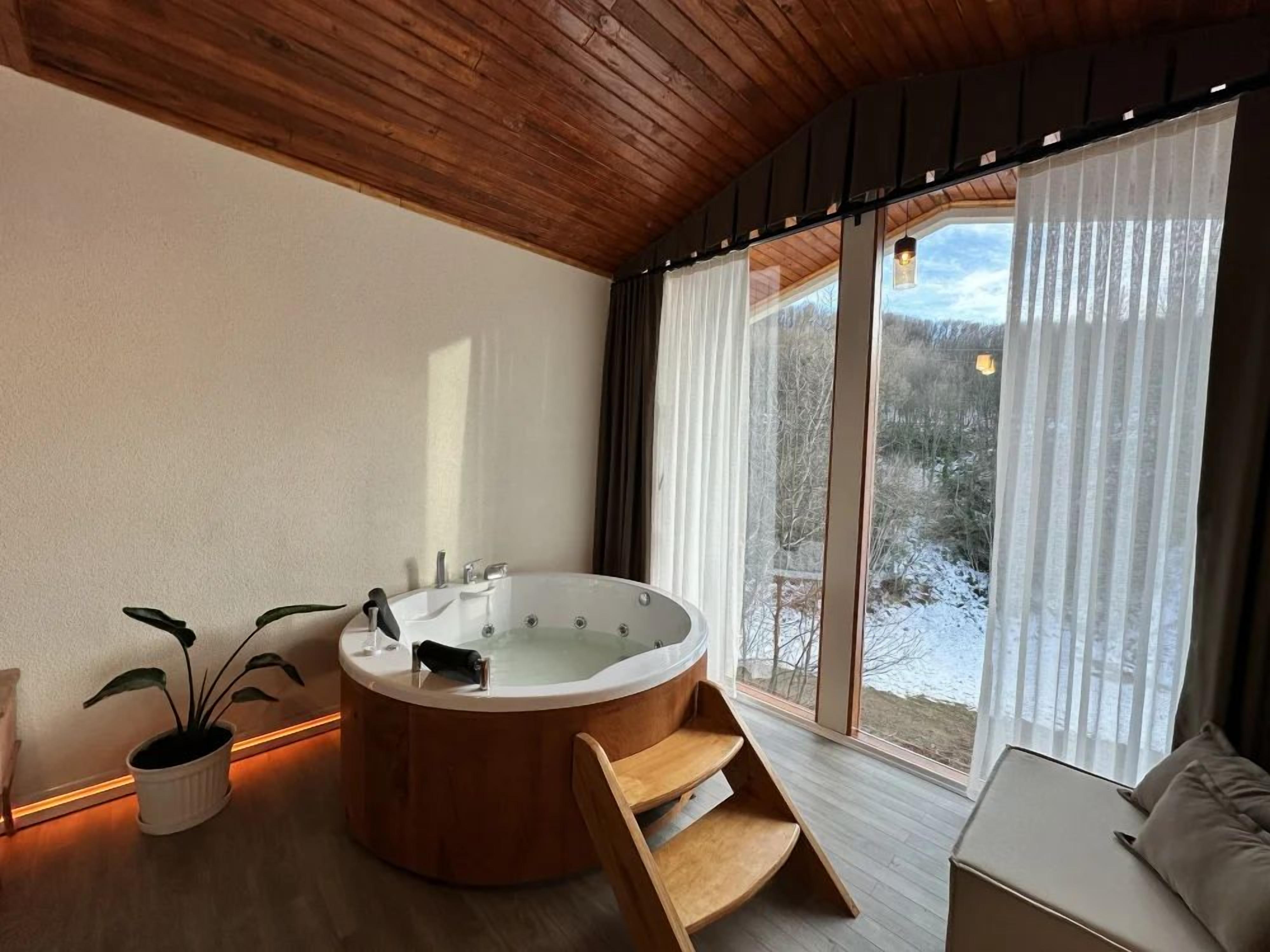 A modern bathroom with a freestanding bathtub, a wooden bench, and a large window with curtains.