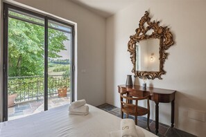 A hotel room with a bed, a desk, a chair, a large mirror, and a view of greenery through a sliding glass door. - Hostdomus - Pool and Wine (Rosignano Monferrato)