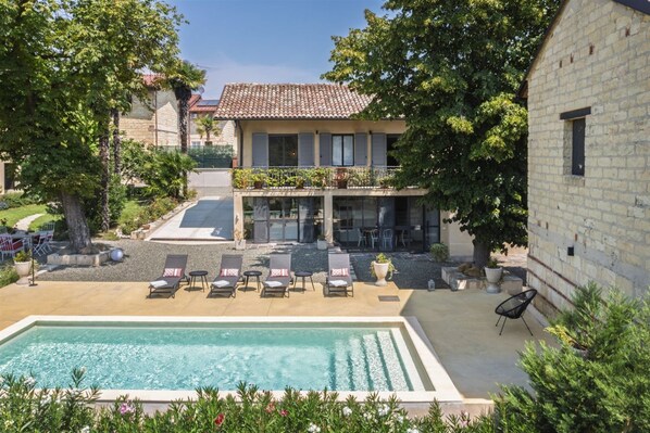 A pool area with lounge chairs, a building with a tiled roof, and a stone wall. - Hostdomus - Pool and Wine (Rosignano Monferrato)