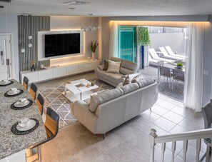 A modern living room with a sectional sofa, a dining area with a table and chairs, a TV mounted on the wall, and a balcony with lounge chairs. - Kissimmee 5BR Modern Home w/ Private Pool (Davenport)