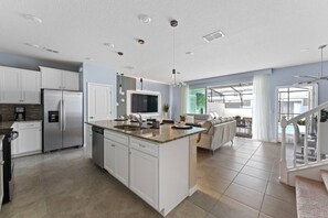A modern kitchen with white cabinetry, a large island, stainless steel appliances, and a dining area with a sofa and table. - Kissimmee 5BR Modern Home w/ Private Pool (Davenport)