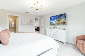 Une chambre à coucher avec un grand lit, une commode blanche, un téléviseur et une porte donnant sur une autre pièce.