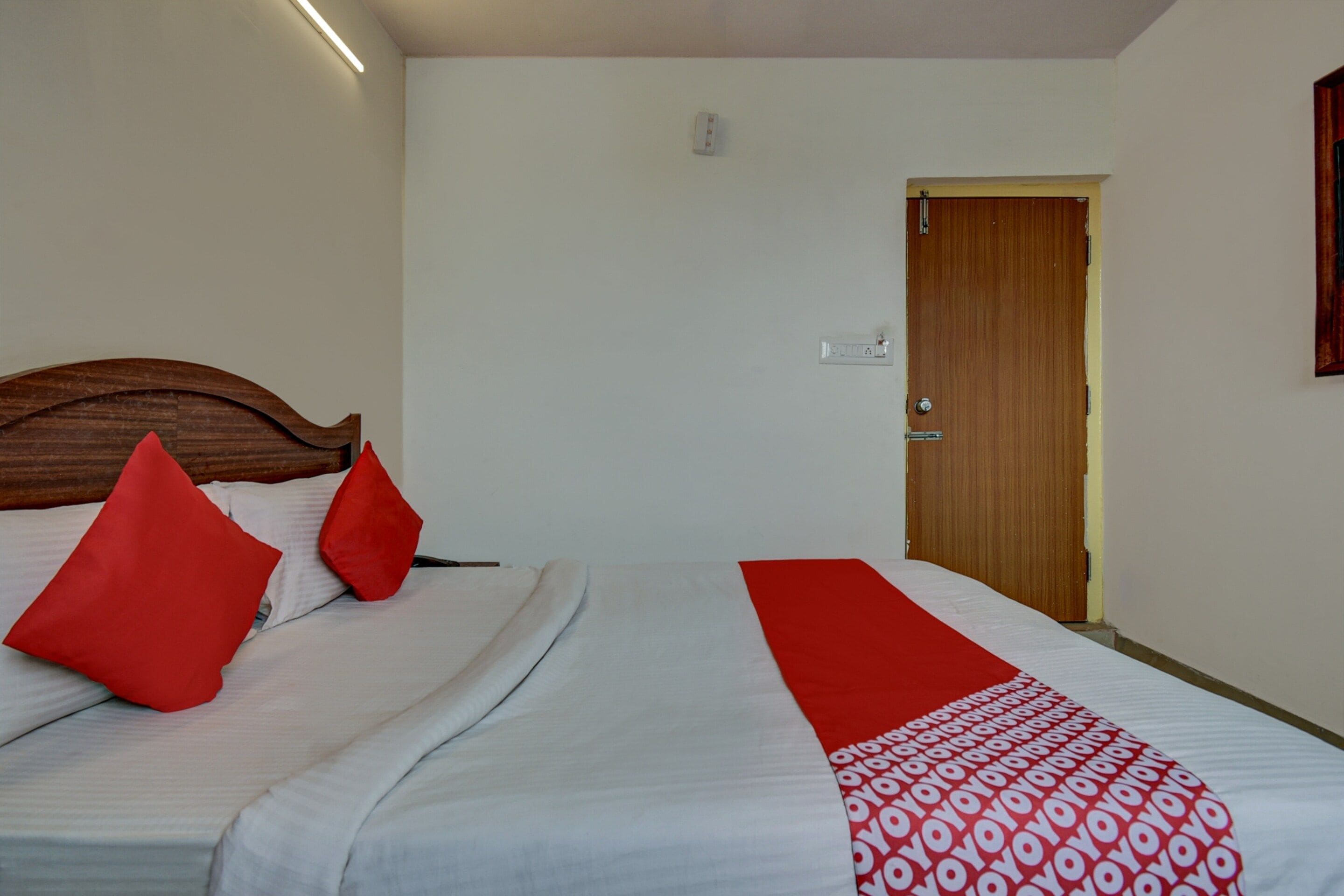 A hotel room with a bed, a wooden headboard, a red throw pillow, a white bedspread, and a red and white patterned blanket.