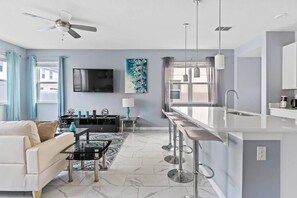 A modern living room with a sofa, a coffee table, and a TV. There is a kitchen area with a bar and a window. - Compass Bay 4bdr Renovated BBQ Near Disney (Kissimmee)