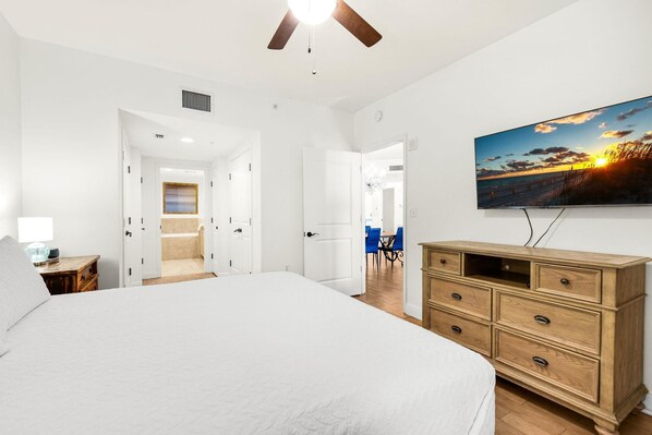 A bedroom with a large bed, a wooden dresser, a TV mounted on the wall, and a bathroom visible through an open door. - Portofino Island Resort 2-1508 (Pensacola Beach)
