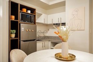 A modern kitchen with a built-in microwave, refrigerator, and stainless steel appliances. There is a dining table with chairs and a vase with dried flowers.