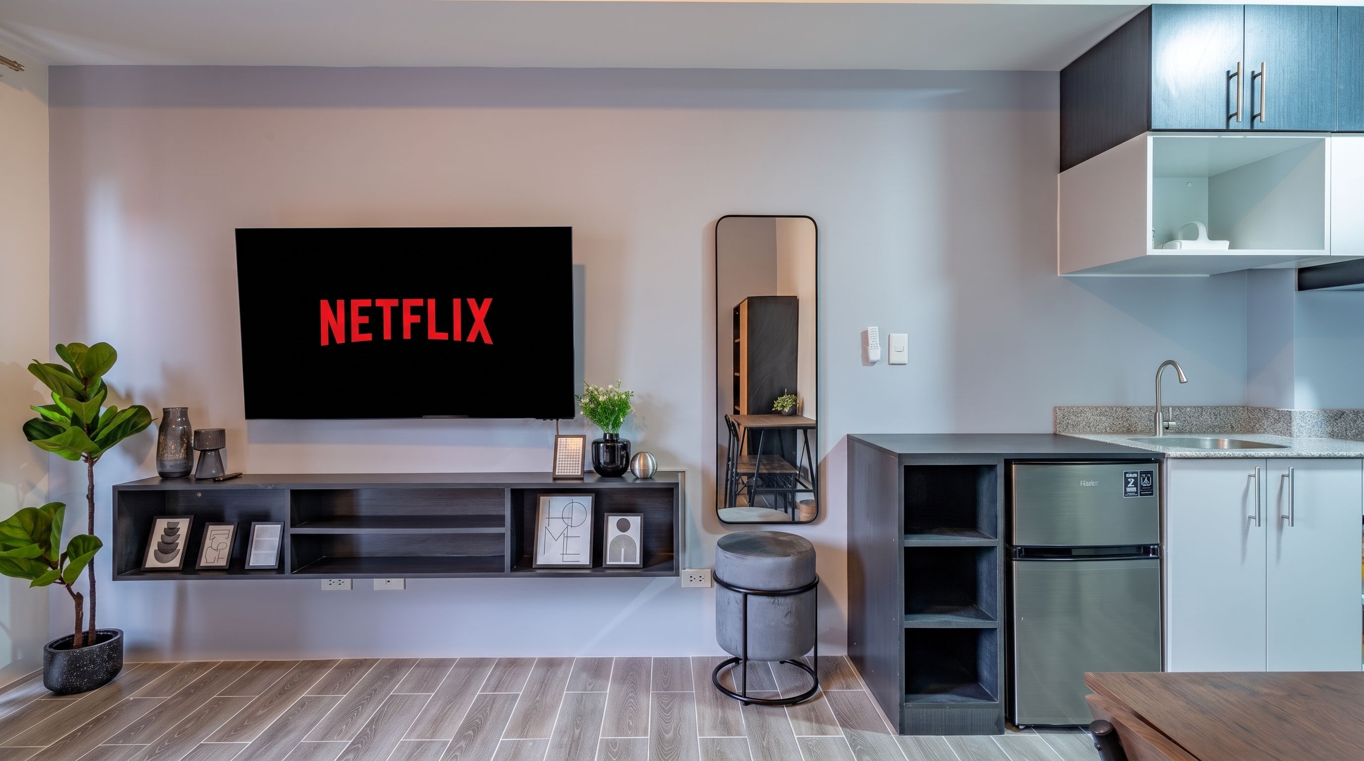 Un salon moderne doté d’un téléviseur à écran plat affichant le logo « NETFLIX », d’une petite plante, de quelques tableaux encadrés, d’un miroir et d’une cuisine minimaliste avec un réfrigérateur et des placards.