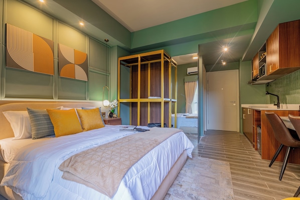 A modern hotel room with a large bed, a wooden wardrobe, a bathroom with a shower, and a kitchenette.