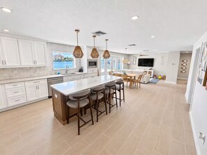 Una cocina moderna con gabinetes blancos, una isla central con taburetes de bar, iluminación colgante y un amplio ventanal con vista a un cuerpo de agua.