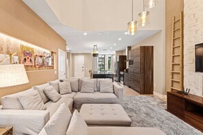 A modern living room with a sectional sofa, a small ottoman, and a TV mounted on a brick-patterned wall. - Magic Village 8guests Jacuzzi 8 min Disney (Kissimmee)