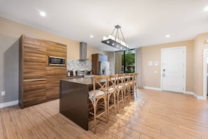 A modern kitchen with a central island, wooden cabinetry, and a dining area with chairs. - 7458 Magic Village 8 Guests - 8 min Disney (Kissimmee)