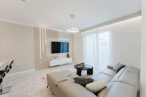 Ein modernes Wohnzimmer mit einem großen Flachbildfernseher, der an einer weißen Konsole befestigt ist, einem beige gefärbten Sofa, einem Couchtisch und einem großen Fenster mit durchsichtigen Vorhängen.