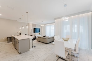 A modern kitchen with a central island, a dining area with a white table and chairs, and a living area with a sofa and a TV. - Storey Lake 4BR Villa With Pool and Disney Rooms (Kissimmee)