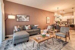 Een moderne woonkamer met een grijze modulaire bank, een salontafel en een eethoek met stoelen, met een keuken op de achtergrond.