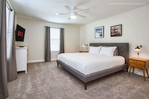 Une chambre à coucher avec un lit, deux tables de chevet avec des lampes, un ventilateur de plafond, un téléviseur posé sur un meuble et une fenêtre avec des rideaux.
