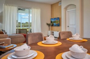 A dining area with a table set for four, a sofa, a painting on the wall, and a view of the outdoors through large windows. - Luxury 2BR / Mickey Room & Resort Amenities! (Davenport)