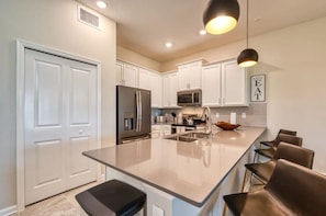 A modern kitchen with white cabinetry, stainless steel appliances, a central island with seating, and pendant lighting. - Luxury 2BR / Mickey Room & Resort Amenities! (Davenport)