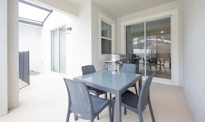 Een moderne eethoek buiten met een glazen tafel en rieten stoelen, een barbecue en een schuifpui naar een terras.
