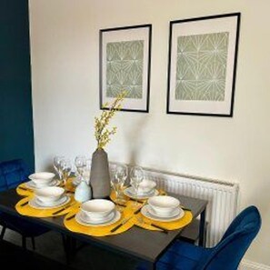 A dining table set with white plates, glasses, and a vase with yellow flowers. There are two framed abstract art pieces on the wall above the table. - Sunderland - Apartment - Stay 4 - Central Location (Sunderland)