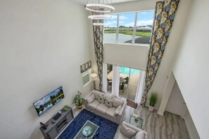 A spacious living area with a high ceiling, a large window overlooking a swimming pool, and a view of a golf course. - 5BR Villa Private Pool & Lake View (Kissimmee)