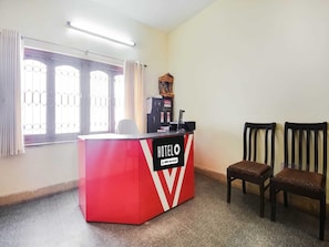 A hotel reception desk with a red and white design, two wooden chairs, a window with metal bars, and a reception counter with a sign. - Deluxe 66 Inn Guest House (Bidhannagar)