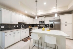 A modern kitchen with white cabinets, a central island with bar stools, stainless steel appliances, and a black tiled backsplash. - Cozy 3BR in Storey Lake 10 Min From Disney! (Kissimmee)