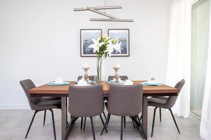 A modern dining room with a rectangular wooden table, six grey chairs, two framed artworks on the wall, and a vase with white flowers. - Luxury 4BR Home w/ Pool Near Disney! (Davenport)