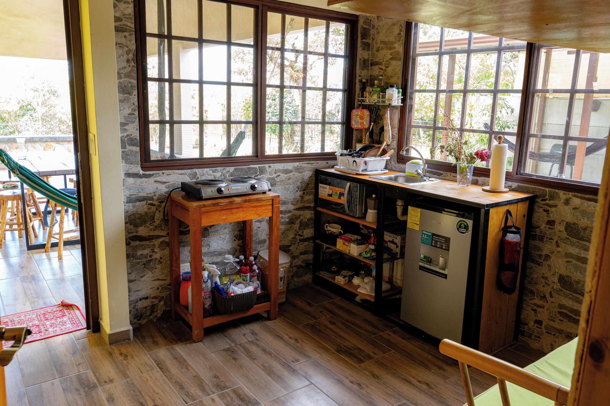 A compact kitchen with a stone wall, wooden cabinets, a small refrigerator, and a window with a view of greenery.