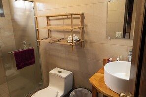 A bathroom with a glass shower enclosure, a white toilet, a wooden shelf with toiletries, and a white sink with a mirror above it. - Your retreat for relaxation in Boquete (Panamá)