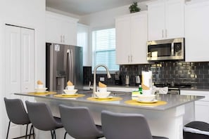 A modern kitchen with white cabinets, a central island with bar stools, stainless steel appliances, and a black tiled backsplash. - Storey Lake 5BR Home Resort + Pool (Kissimmee)