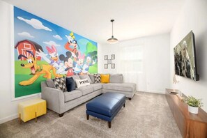 A living room with a large cartoon mural, a grey sectional sofa, a blue ottoman, a yellow radiator, and a TV on the wall. - Luxury 6BR Home at Storey Lake Near Disney (Kissimmee)