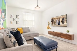 A modern living room with a grey sectional sofa, a blue ottoman, a wooden console table, and a large framed picture on the wall. - Luxury 6BR Home at Storey Lake Near Disney (Kissimmee)