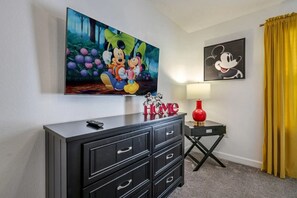 A room with a large Disney-themed painting, a smaller Mickey Mouse painting, a black console table with drawers, a red lamp, and a yellow curtain. - Stylish Home With Private Pool 10 Min From Disney (Kissimmee)