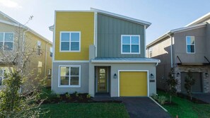 A two-story residential building with a yellow garage door, a grey upper floor, and a front entrance with a small porch. - 5BR Villa w/ Pool & Disney Themes (Kissimmee)
