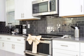 A modern kitchen with white cabinets, a stainless steel microwave, a black tiled backsplash, and a towel with a bow on the oven handle. - 5BR Villa w/ Pool & Disney Themes (Kissimmee)